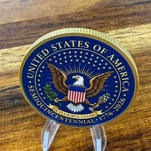 May include: A commemorative coin with a gold rim and a blue center featuring the United States of America seal. The seal includes an eagle, stars, and the text "Semiquincentennial 1776-2026". The coin is displayed on a clear acrylic stand.