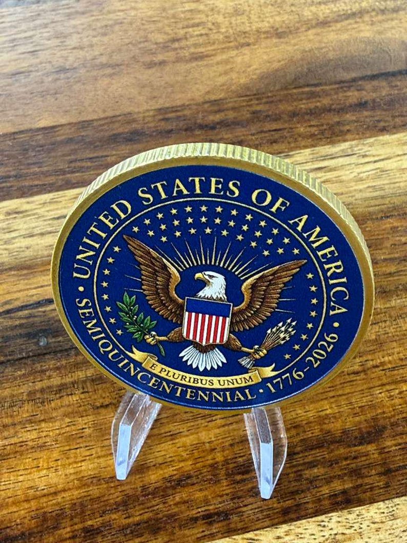May include: A commemorative coin with a gold rim and a blue center featuring the United States of America seal. The seal includes an eagle, stars, and the text "Semiquincentennial 1776-2026". The coin is displayed on a clear acrylic stand.