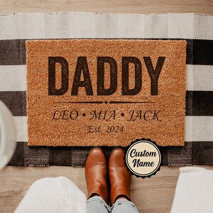 May include: A rectangular doormat with the word "DADDY" in large, dark letters. Below, it reads "LEO • MIA • JACK Est. 2024". The mat is brown, with a black and white checkered rug underneath. Brown boots are visible.