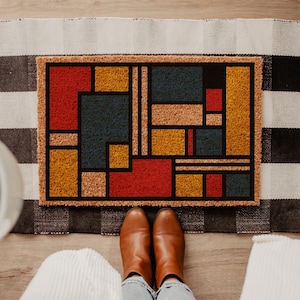 May include: A rectangular doormat with a geometric design in red, yellow, blue, and tan. The design is framed by a black border and set against a black and white checkered rug. Brown boots and blue jeans are visible.