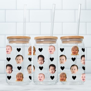 May include: Three clear glass tumblers with bamboo lids and glass straws. Each tumbler is decorated with black heart shapes and images of babies. The tumblers are on a white surface against a white brick wall.