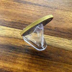 May include: A gold-colored coin with a detailed design, displayed on a clear acrylic stand. The coin has a textured edge and a dark blue center. The stand is angled, showcasing the coin's side profile.