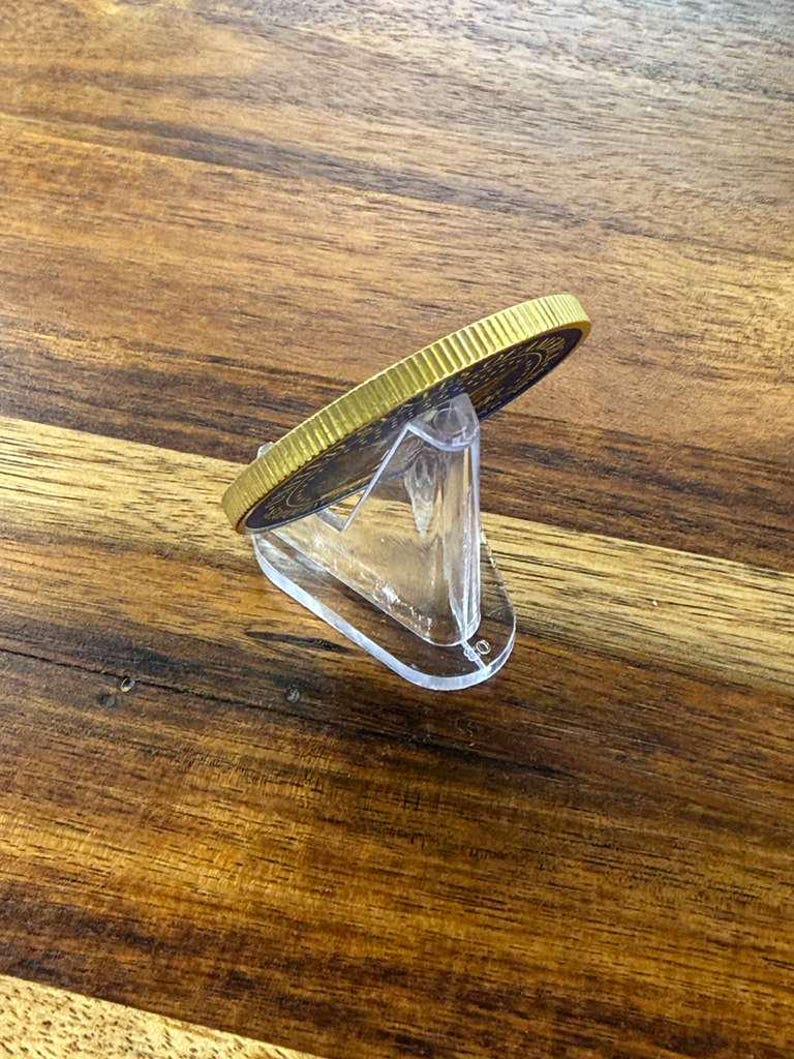 May include: A gold-colored coin with a detailed design, displayed on a clear acrylic stand. The coin has a textured edge and a dark blue center. The stand is angled, showcasing the coin's side profile.