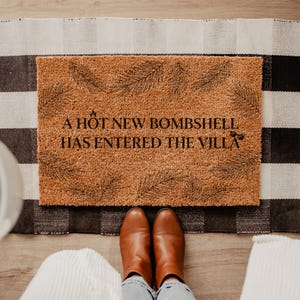 May include: A rectangular doormat with the text "A HOT NEW BOMBSHELL HAS ENTERED THE VILLA" in black. The mat is brown with a textured surface and a fern leaf design. The mat is on a black and white checkered rug.