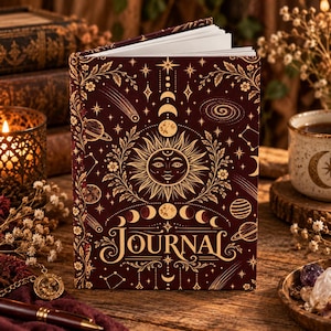 May include: A hardbound journal with a deep burgundy cover featuring gold foil celestial illustrations of a sun face, moon phases, stars, and planets. The word JOURNAL is printed in a decorative gold font at the bottom center of the front cover.