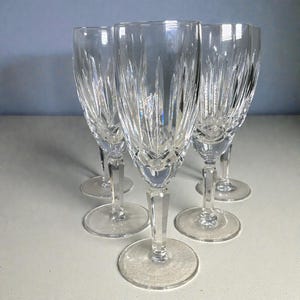 May include: A set of five clear crystal wine glasses. Each glass features a detailed, etched pattern on the bowl and a faceted stem. The glasses are arranged on a light-colored surface, showcasing their elegant design.