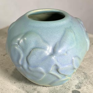 May include: A spherical ceramic vase with a light blue, iridescent glaze. The surface features raised, organic, cloud-like relief patterns that create a textured appearance. The vase has a circular opening at the top and a smooth, rounded body.