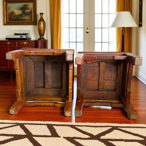 May include: Two antique wooden stools, viewed from below, showcasing their aged construction. The stools are a warm brown color, with visible wood grain and joinery. The legs are sturdy and angled, suggesting a traditional design. The stools are placed on a hardwood floor.