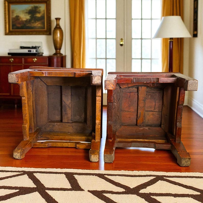 May include: Two antique wooden stools, viewed from below, showcasing their aged construction. The stools are a warm brown color, with visible wood grain and joinery. The legs are sturdy and angled, suggesting a traditional design. The stools are placed on a hardwood floor.