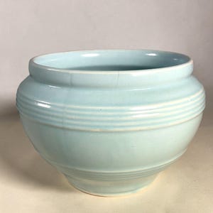 May include: A light blue ceramic bowl with a glossy finish and a rounded, bulbous shape. The exterior features three horizontal ridges encircling the upper section, with a smooth, curved base and a wide, open rim at the top of the vessel.