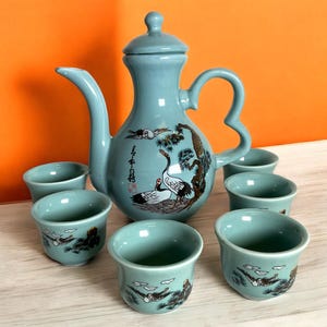 May include: A teal blue ceramic tea set with a teapot and six cups. The teapot has a lid and a handle. The cups are small and have a rounded shape. The set is decorated with a design of cranes and pine trees. The teapot has Chinese characters on it.