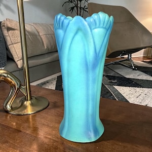 May include: A tall, turquoise-colored vase with a flared base and a scalloped rim. The vase has a smooth, glossy finish and a subtle, swirling pattern of lighter blue. The vase is likely made of glass or ceramic and is designed for displaying flowers.