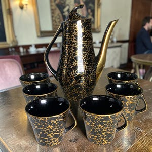 May include: A ceramic coffee set featuring a tall lidded pitcher and five matching cups. Each piece has a glossy black base with a textured gold splatter pattern. The pitcher has a curved handle and a long spout, while the cups have handles.