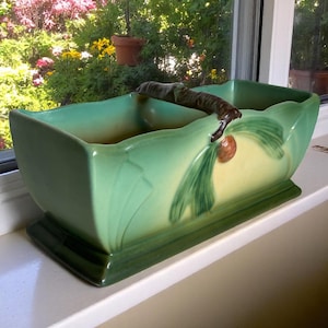 May include: A rectangular, green ceramic planter with a handle. The planter has a light green and yellow interior, with a dark green base. The handle is brown and the planter is decorated with green leaves and a brown pinecone.