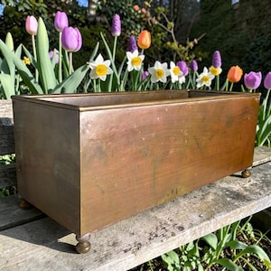 May include: A rectangular copper planter box with a smooth, weathered finish and a metallic sheen. The container features a simple, clean-lined design with four small, rounded ball-shaped feet at the base, providing a stable foundation.