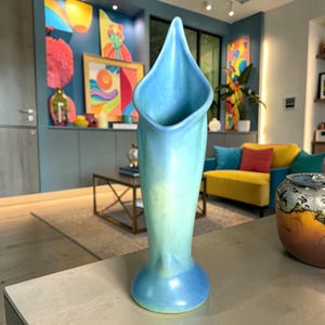 May include: A tall, light blue ceramic vase with a flared, petal-like opening. The vase has a rounded base and a smooth, gradient finish, transitioning from a darker blue at the base to a lighter blue at the top. The vase is displayed on a light-colored surface.