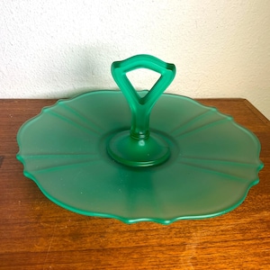 May include: A green glass serving tray with a handle. The tray has a scalloped edge and a frosted finish. The handle is a triangular shape. The tray is sitting on a wooden surface.