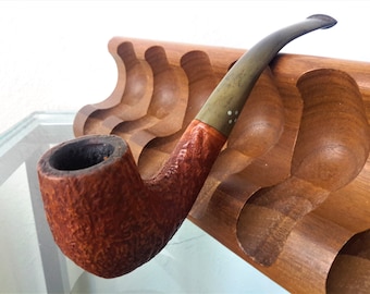 May include: A tobacco pipe with a textured, reddish-brown bowl and a curved, brass-colored stem. The pipe rests in a wooden pipe rack. The stem has a brass band with white dots. The pipe is a classic smoking accessory.