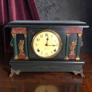 May include: A black mantel clock with a cream-colored face and the text Sessions. The clock features green and red marble-effect columns, gold-toned decorative figures, and ornate metal feet. The dial displays black Arabic numerals and hands.