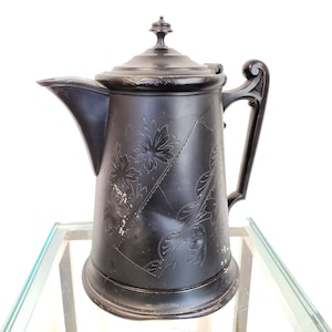 May include: A matte black teapot featuring a tapered cylindrical body with an integrated handle and a spout. The exterior surface is decorated with etched floral patterns and geometric lines. A matching lid with a small knob sits on top.