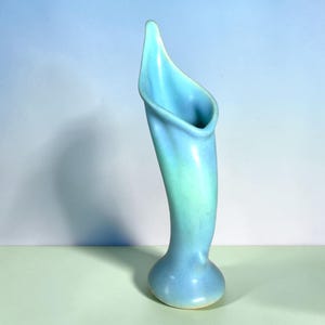 May include: A tall, slender ceramic vase with a smooth, matte finish. The object features a gradient color scheme, transitioning from a soft sky blue at the top to a lighter, pale turquoise hue toward the rounded, stable base. The opening is elongated.