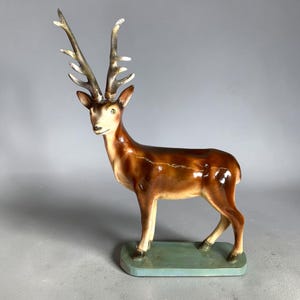 May include: A ceramic deer figurine featuring a glossy brown coat, a cream-colored face, and tall, textured antlers. The animal stands on a rectangular, light blue base, with its head turned slightly to the left, showcasing a smooth, glazed finish.