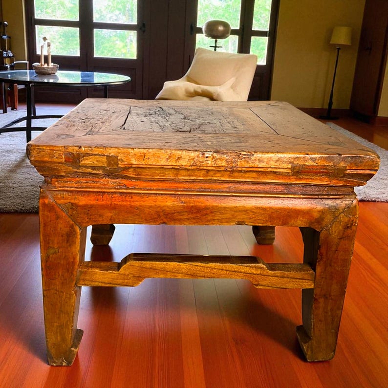 May include: A rectangular, antique wooden coffee table with a distressed finish. The table has a warm, reddish-brown hue with visible wood grain and unique joinery details. The legs are sturdy and feature decorative cutouts, showcasing a vintage aesthetic.