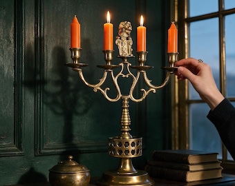May include: A brass candelabra featuring four candle holders with lit orange taper candles and a central decorative lion emblem. The metal stand has a tiered base with a lattice pattern and a matching small brass lidded container.