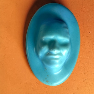 May include: A light blue, oval-shaped wall hanging featuring a sculpted face. The face is centered within the oval, with a smooth, matte finish. The wall hanging is mounted on an orange wall.