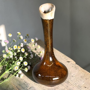 May include: A tall, brown ceramic vase with a flared, off-white rim. The vase has a slender neck and a rounded base. The surface has a glossy finish with a speckled, metallic appearance. Daisies with white and yellow petals are in the background.