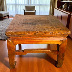 May include: A rectangular, low coffee table with a distressed, aged wood finish. The table has a thick top and a sturdy, carved base. The wood tones range from light to dark brown, with visible grain and wear. The table is approximately 48 inches long, 30 inches wide, and 16 inches high.