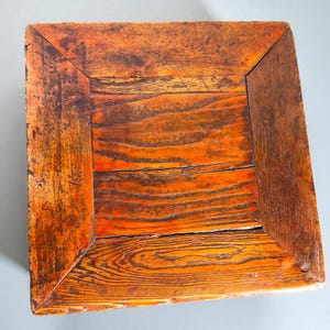May include: A square, wooden table top with a distressed, orange-brown finish. The wood grain is visible, and the corners are darker. The table top has a rustic, vintage appearance.