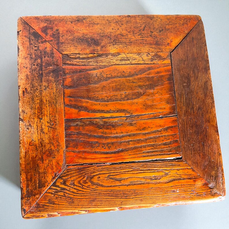 May include: A square, wooden table top with a distressed, orange-brown finish. The wood grain is visible, and the corners are darker. The table top has a rustic, vintage appearance.