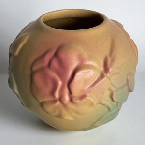 May include: A round, peach-colored ceramic vase with a wide opening. The vase features a raised floral design with accents of pink and green. The overall shape is spherical, and the surface has a matte finish. This decorative vase is suitable for home decor.