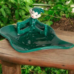 May include: A ceramic figurine of a green elf sitting on a leaf-shaped dish. The elf has a green outfit, pointed ears, and a red mouth. The dish and elf are a glossy, dark green color. The figurine is placed on a wooden surface.