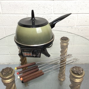 May include: A vintage green and black electric fondue pot with a black handle and a round base. The pot is sitting on a glass table with four wooden skewers with red, yellow, and orange tips.
