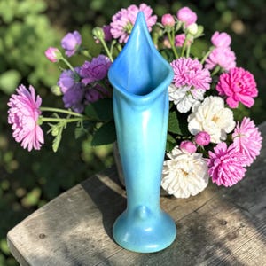 May include: A tall, slender ceramic vase with a smooth, matte finish in a gradient of light blue to turquoise. The vase features a flared, pointed rim that tapers down to a narrow neck and a circular base, standing upright with a soft sheen.