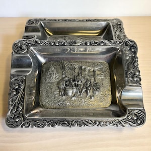 May include: Two vintage silver-toned ashtrays with ornate, scrolled edges. The center of each tray features a rectangular, raised relief with a detailed scene of people gathered around a barrel. The ashtrays are rectangular with rounded corners.