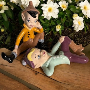 May include: Two ceramic elf figurines. One is seated, wearing a yellow jacket, brown pants, and black boots. The other is reclining, dressed in a light green top and purple pants. Both have painted faces and are surrounded by white flowers.