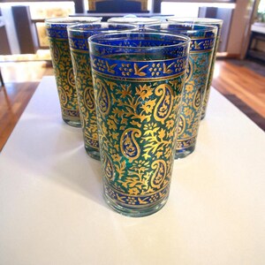 May include: Set of six tall drinking glasses. Each glass features a teal and gold paisley design with a blue band at the top and bottom. The glasses are arranged on a white surface.