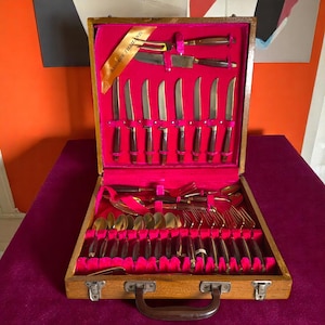 May include: A wooden carrying case with a red velvet interior holds a set of gold-toned silverware. The case has a brass handle and a label that reads "BANGKOK THAILAND".