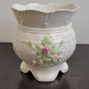 May include: Cream-colored ceramic planter with a scalloped rim and floral embellishments. The planter features a raised floral design with a pink rose, green leaves, and small blue and pink flowers. It has a rounded body and sits on three decorative feet.