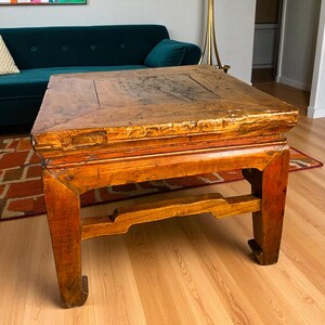 May include: A square, antique wooden coffee table with a distressed finish. The table has a warm, reddish-brown hue and features a sturdy, rectangular base with decorative cutouts. The tabletop shows signs of wear, adding to its vintage charm.