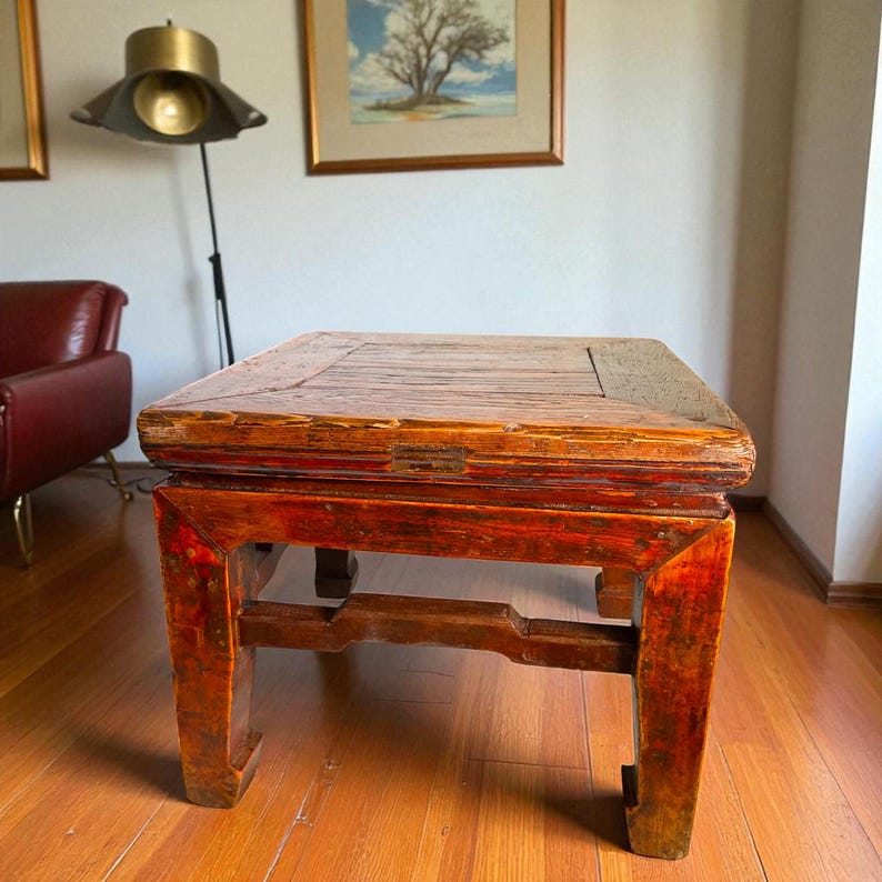 May include: A rectangular, antique wooden coffee table with a distressed, reddish-brown finish. The table has a square top and sturdy legs, showcasing traditional joinery. The wood grain is visible, adding to its vintage appeal. The table measures approximately 30 inches long, 30 inches wide, and 18 inches high.