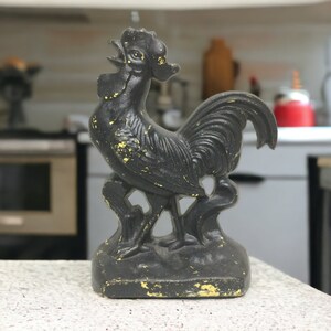 May include: Black cast iron rooster doorstop with a yellowed patina. The rooster is standing on a base with a leafy design.