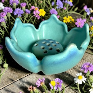 May include: A turquoise ceramic flower frog with a central, perforated sphere, set within a petal-shaped bowl. The frog is surrounded by colorful flowers, creating a vibrant contrast against the wooden surface.