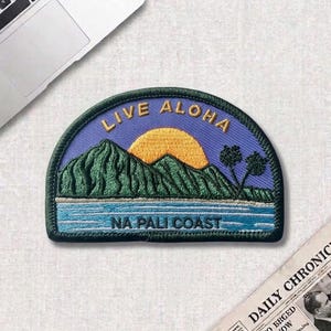 May include: Embroidered patch with a scenic design featuring mountains, a setting sun, and ocean waves. The patch has the words "LIVE ALOHA" and "NA PALI COAST" in gold and white lettering against a blue background.