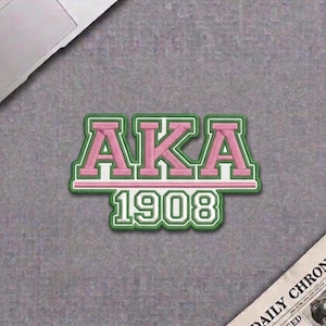May include: Embroidered patch featuring the letters "AKA" in pink, outlined in white and green, with the year "1908" below. The patch is on a gray surface, with a laptop and newspaper in the background.
