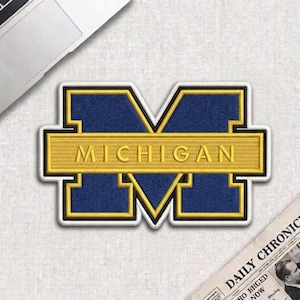 May include: Embroidered patch featuring the letter "M" in navy blue with a gold border and the word "MICHIGAN" in gold. The patch is on a white surface, with a laptop and newspaper in the background.