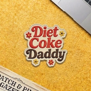 May include: Embroidered patch with the words "Diet Coke Daddy" in red and brown, with floral accents. The patch is on a yellow textured surface, with a laptop and a newspaper visible in the background.